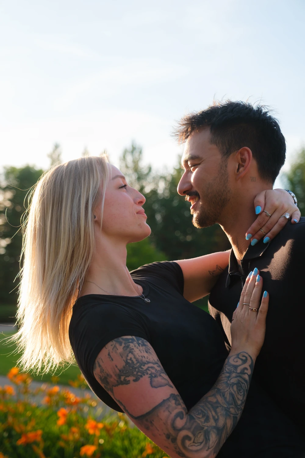 A couple embracing and looking at one another in warm sunlight, with green foliage and flowers behind them.