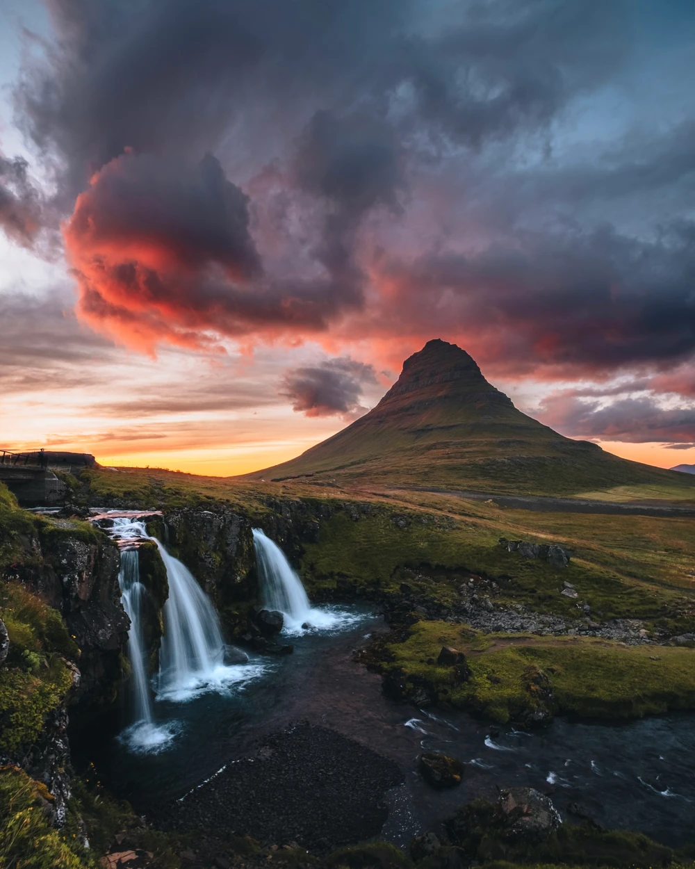 A waterfall beneath a pointed mountain and vivid sunset clouds in Iceland.