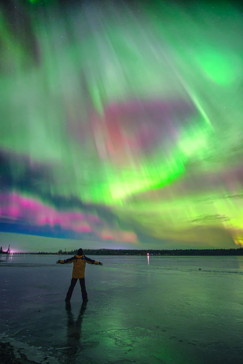 A person with outstretched arms beneath a sweeping green and pink aurora.
