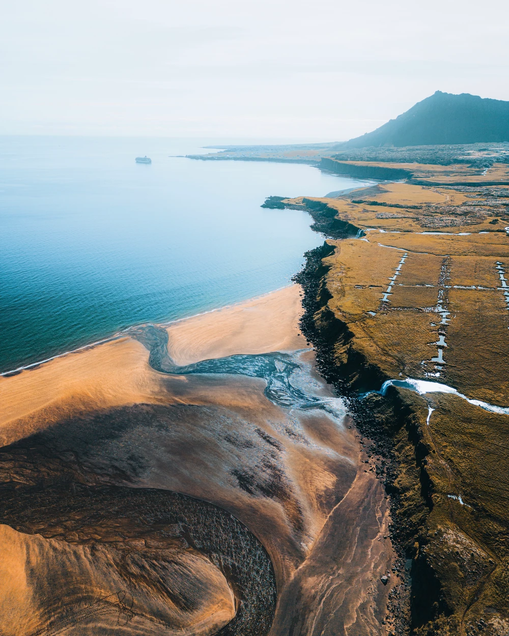 An aerial view of blue water, golden coastal land and dark tidal patterns.