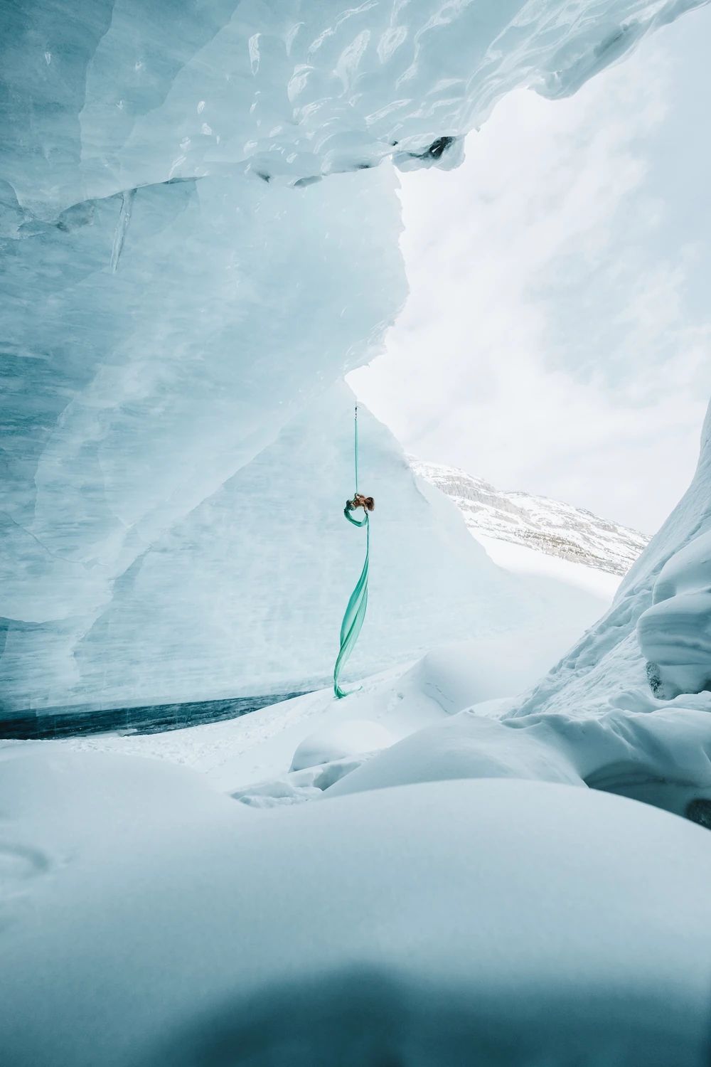 A famous aerial performer, Sasha Galitzki suspended on green silk inside an immense blue ice formation.