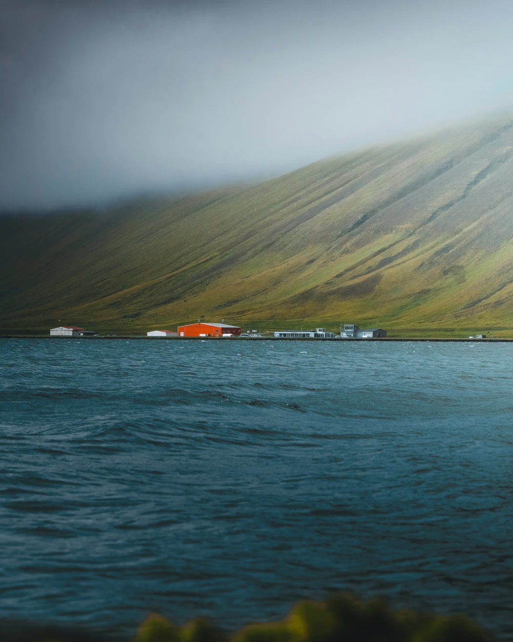 A small orange building beneath a mist-covered mountainside across blue water.