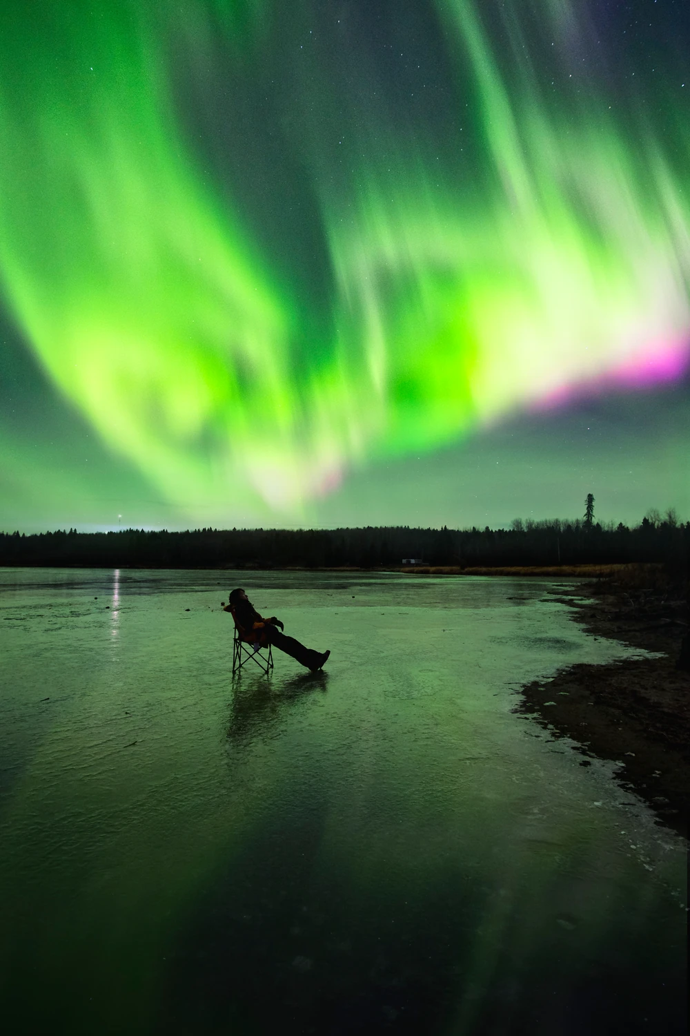 A seated silhouette beneath vivid green and pink northern lights reflected in a lake.