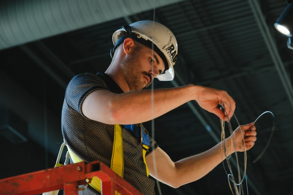 An installer in a hard hat and harness working with cables.