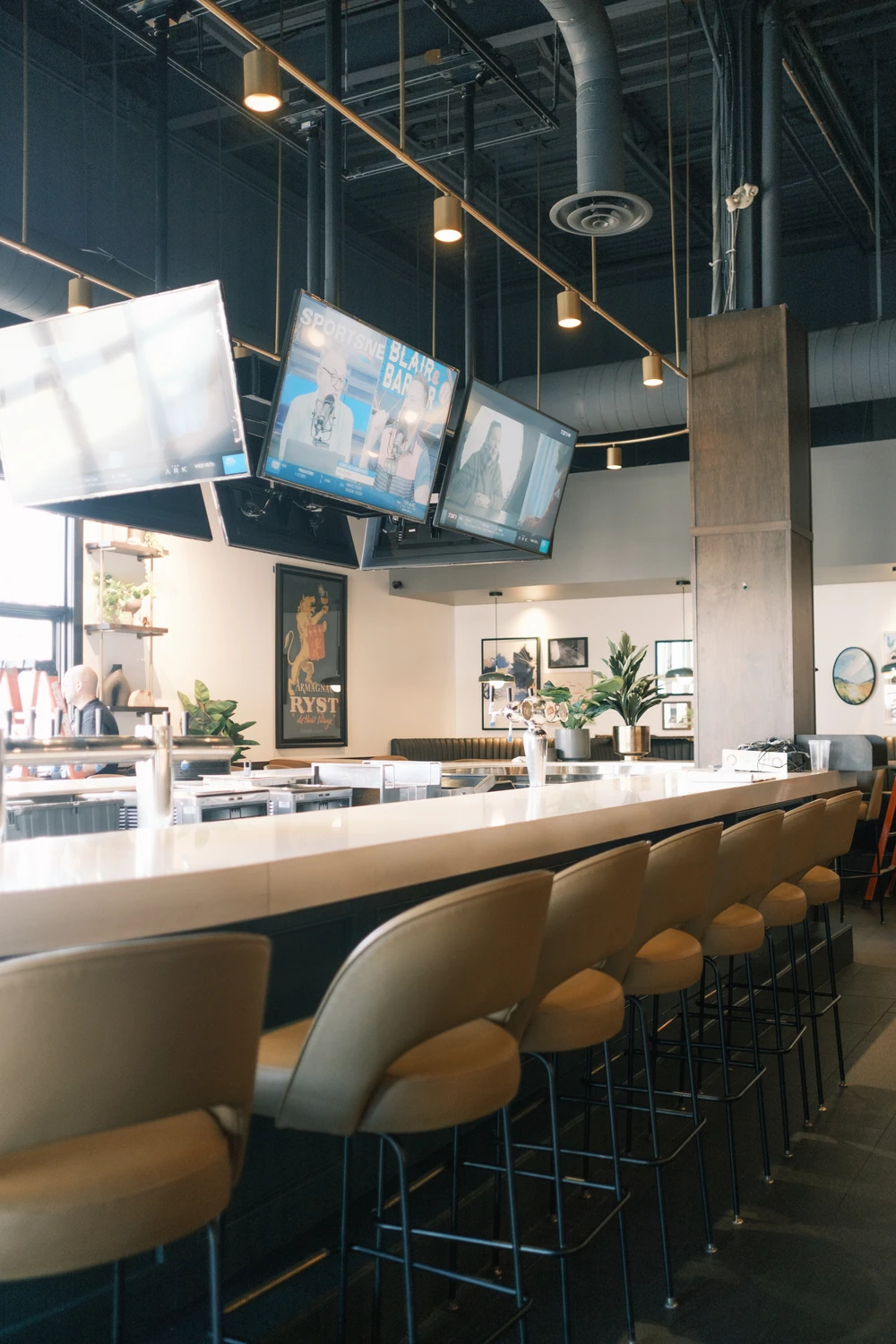 A long pale bar counter and tan stools beneath hanging screens and pendant lights, with framed artwork at Earls.