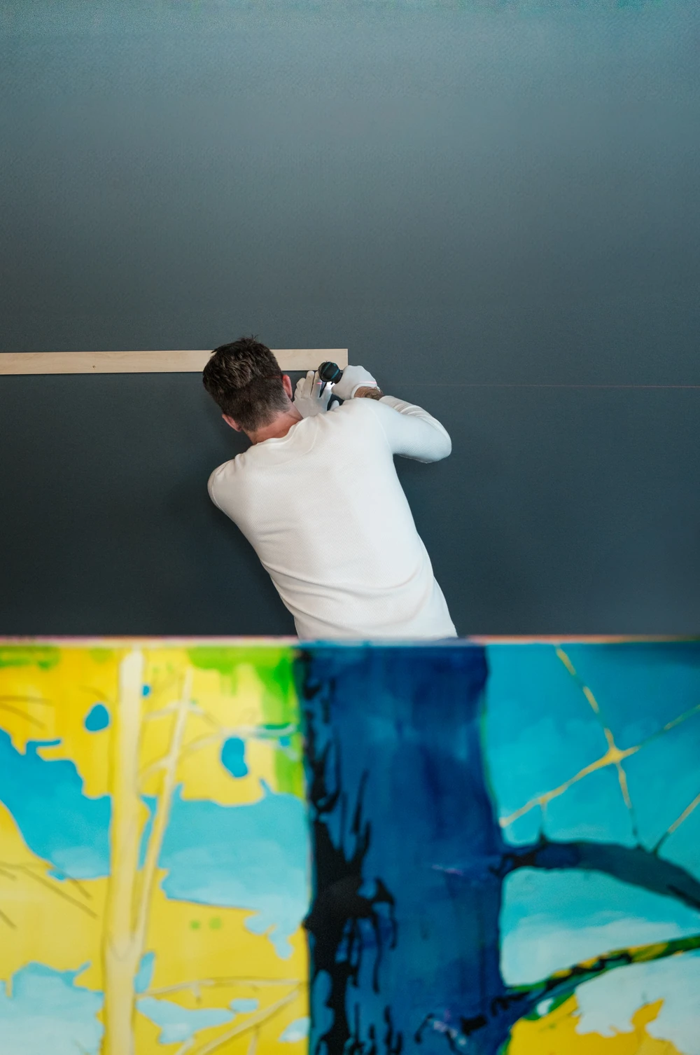 An installer using a drill on a dark wall above a vivid blue and yellow painting at Cross Cancer.