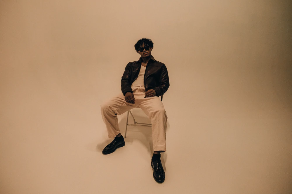An artist seated against a warm studio backdrop in a dark jacket, cream trousers and black boots.