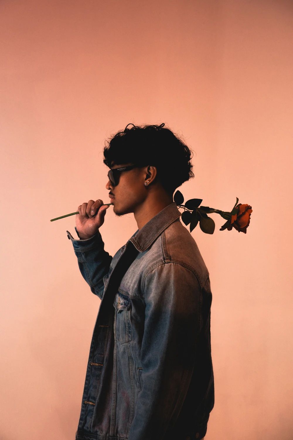 An artist in profile holding a rose against a warm orange studio backdrop.