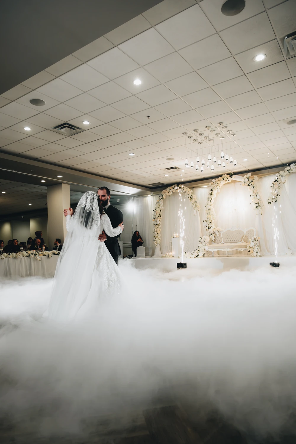 A wedding couple dancing amid low-lying fog in a reception room decorated with white flowers.