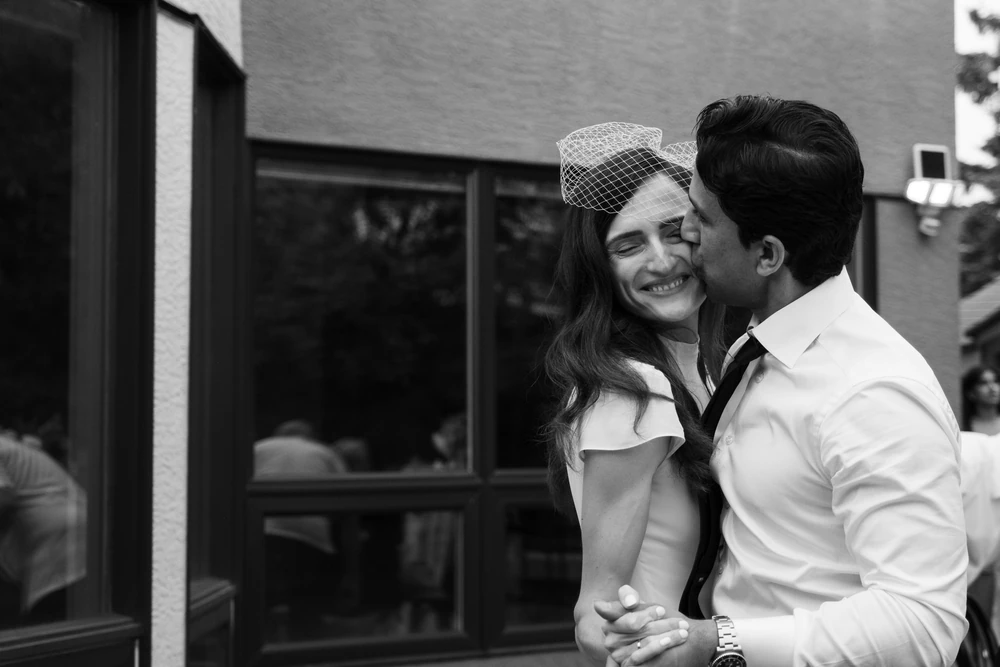 A black-and-white photograph of a bride smiling as her partner kisses her cheek.