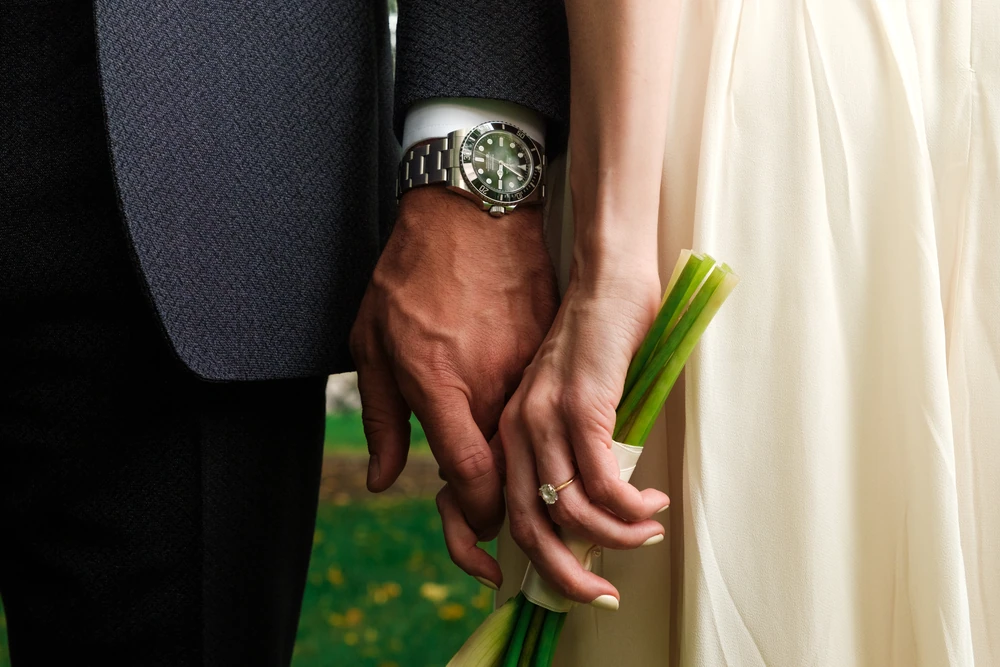A close view of wedding rings, joined hands, a watch and bouquet.