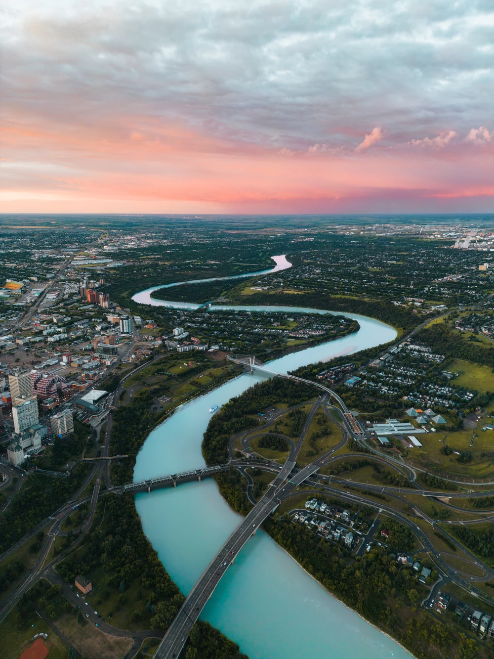 Edmonton’s river valley and bridges winding beneath a pink evening sky, seen from above.
