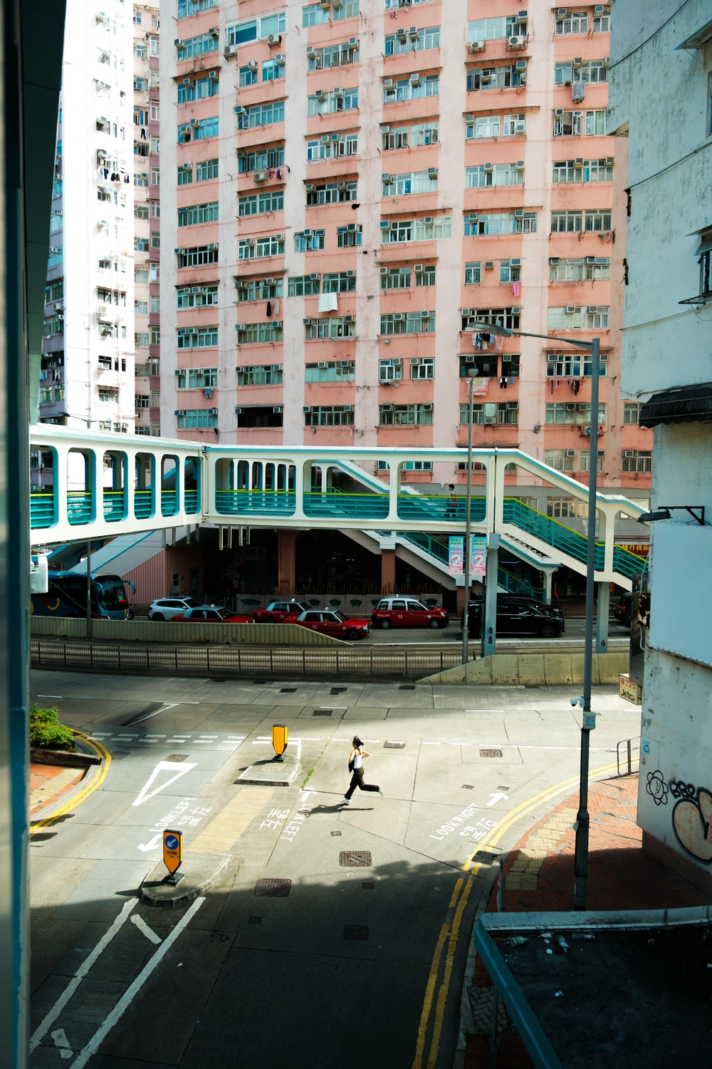 A pedestrian crossing a sunlit Hong Kong street below a footbridge and pink apartment building.