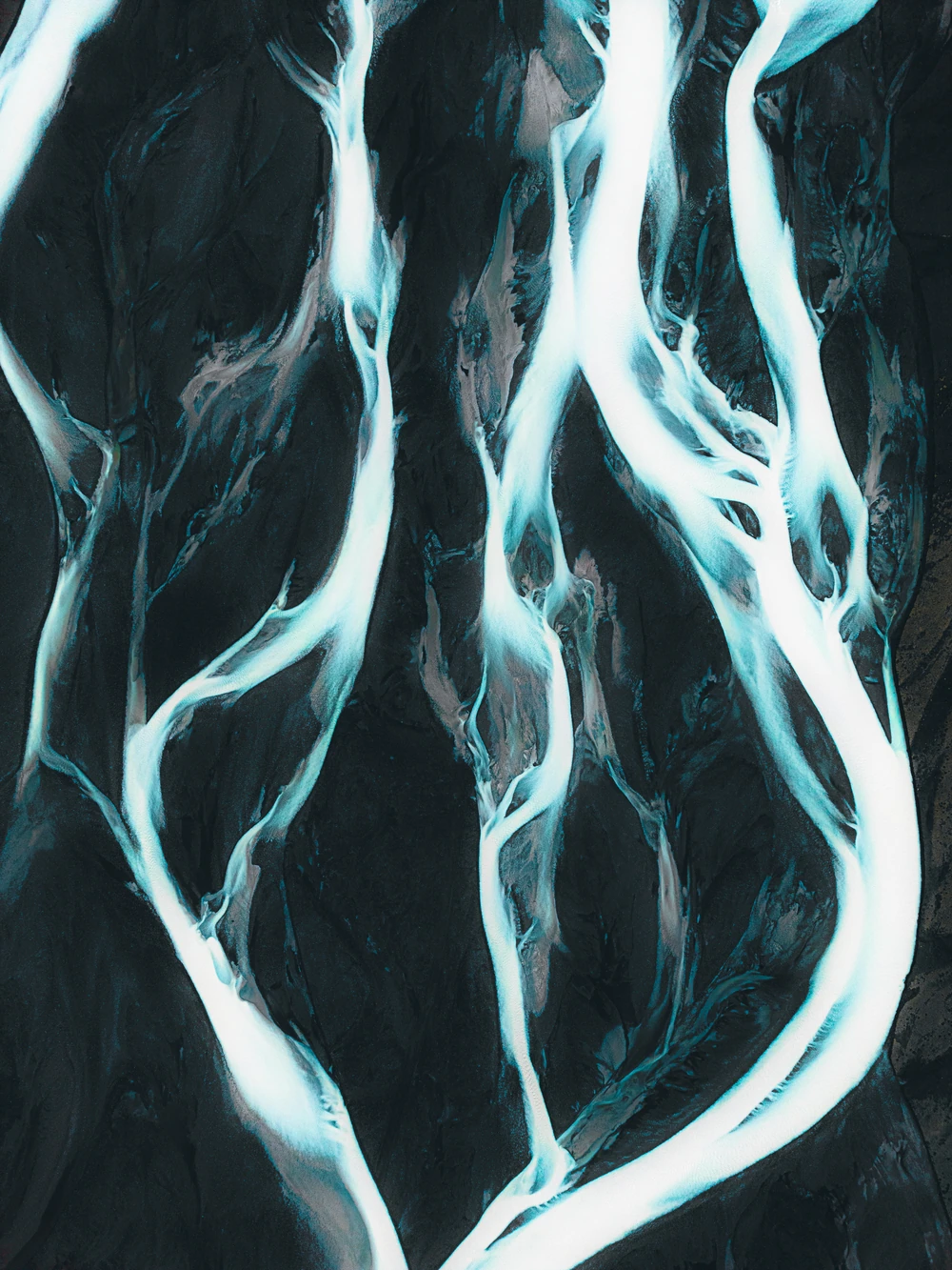 Pale turquoise river channels branching across dark volcanic ground in an abstract aerial view.