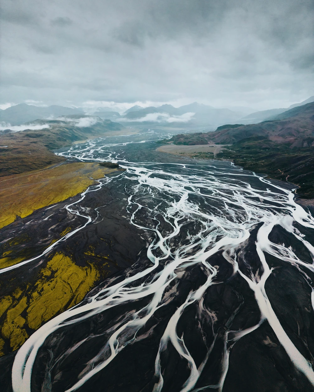 A broad network of pale river channels across dark Iceland terrain beneath misty mountains.