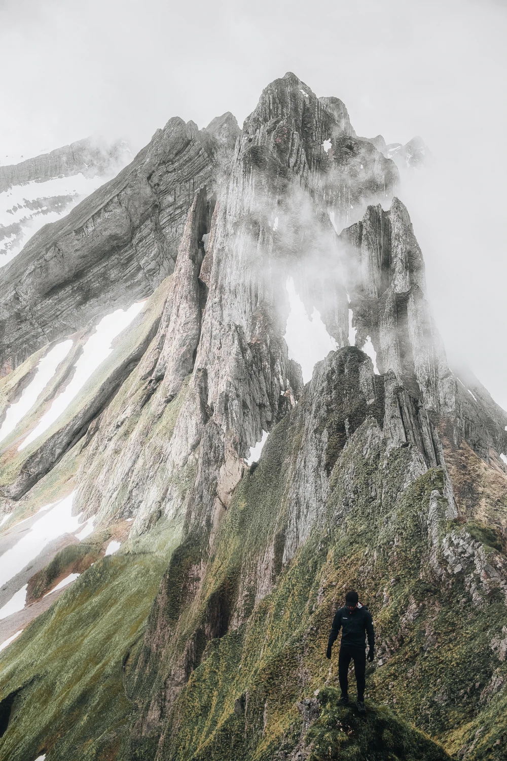A lone hiker standing on a grassy ridge beneath steep rock peaks and passing cloud.
