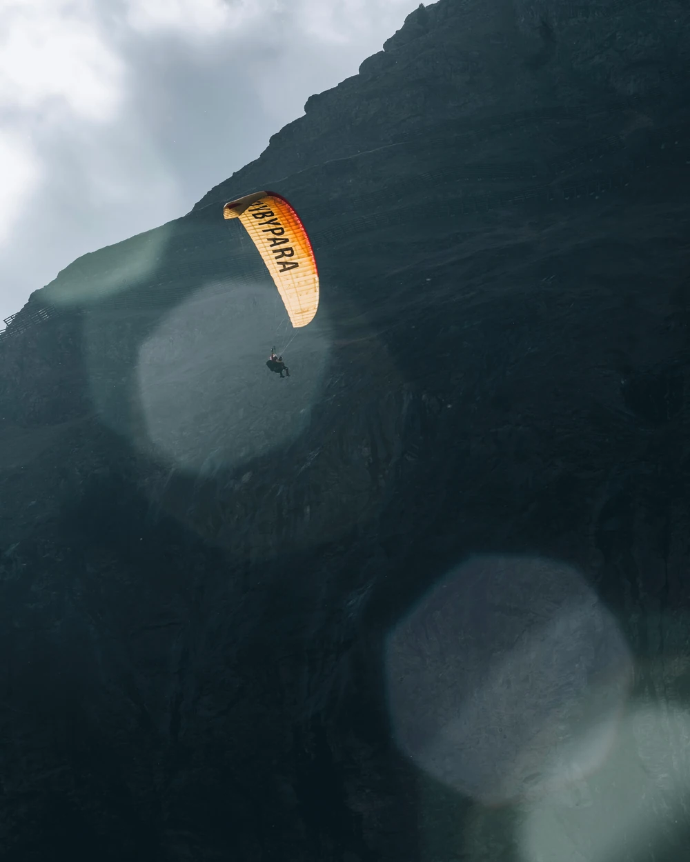A yellow paraglider against a dark mountain wall, seen through soft foreground reflections.