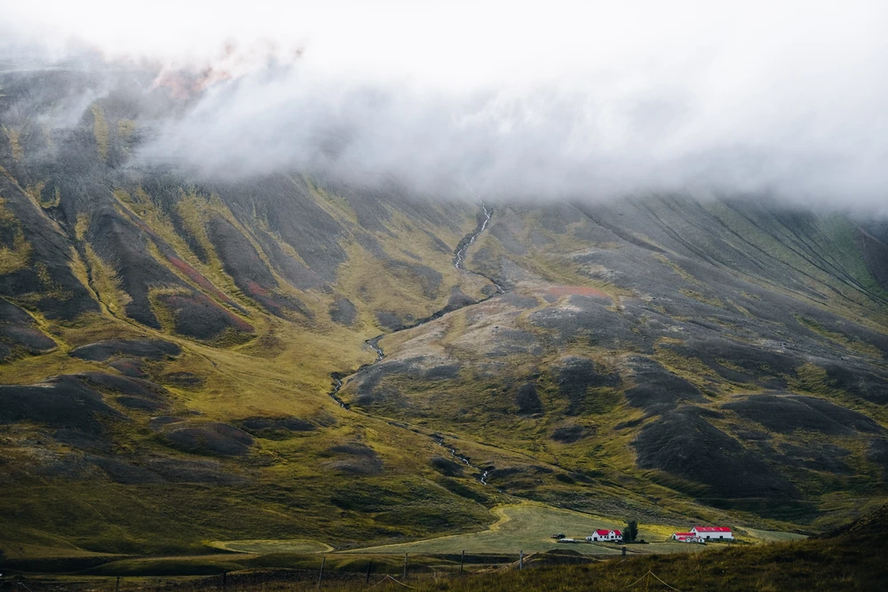 Small red-roofed buildings beneath a vast green mountainside disappearing into cloud.