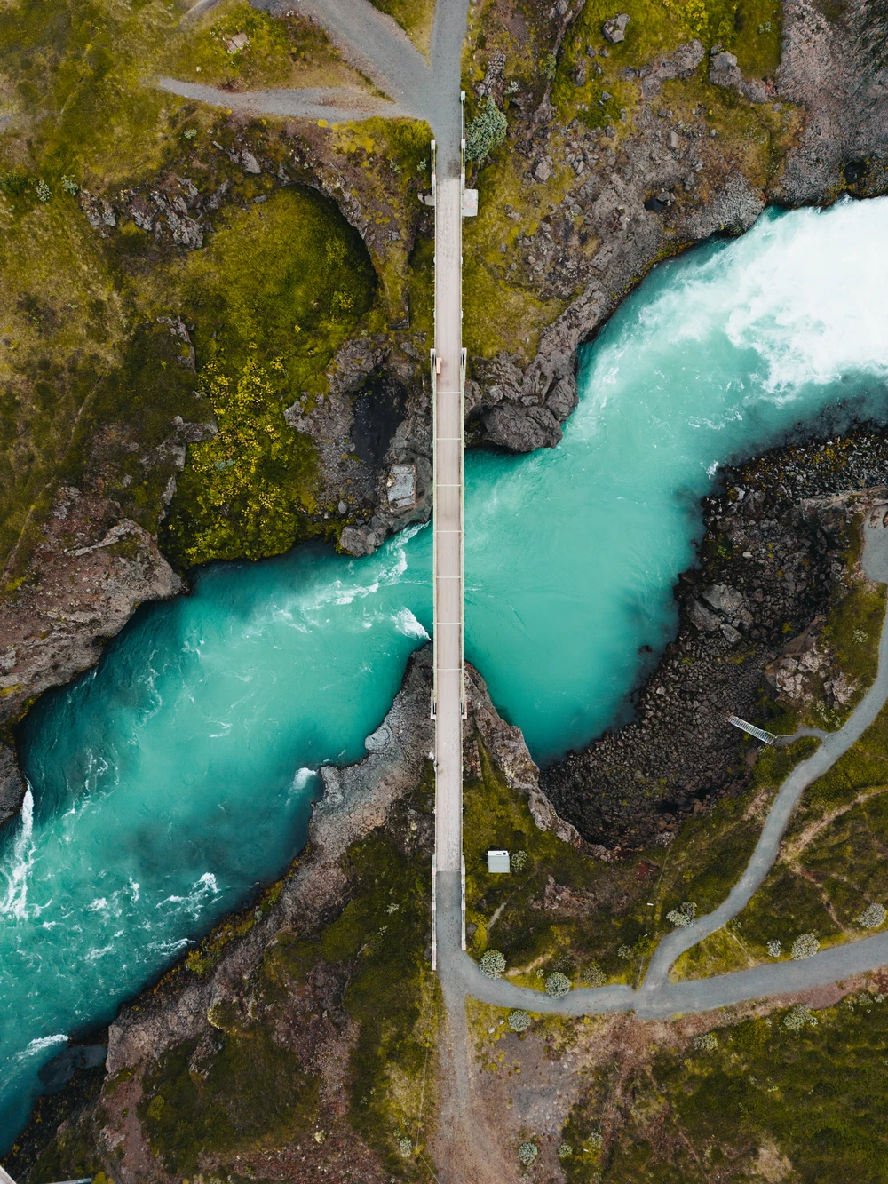 An overhead view of a narrow footbridge crossing a bright turquoise river through rocky ground.