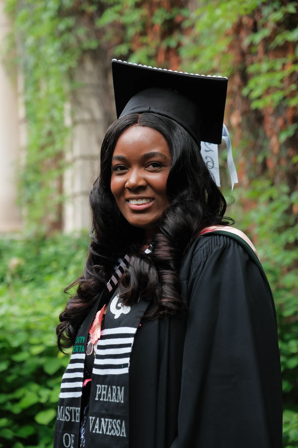 A smiling graduate in a cap and gown beside green ivy and stone columns.