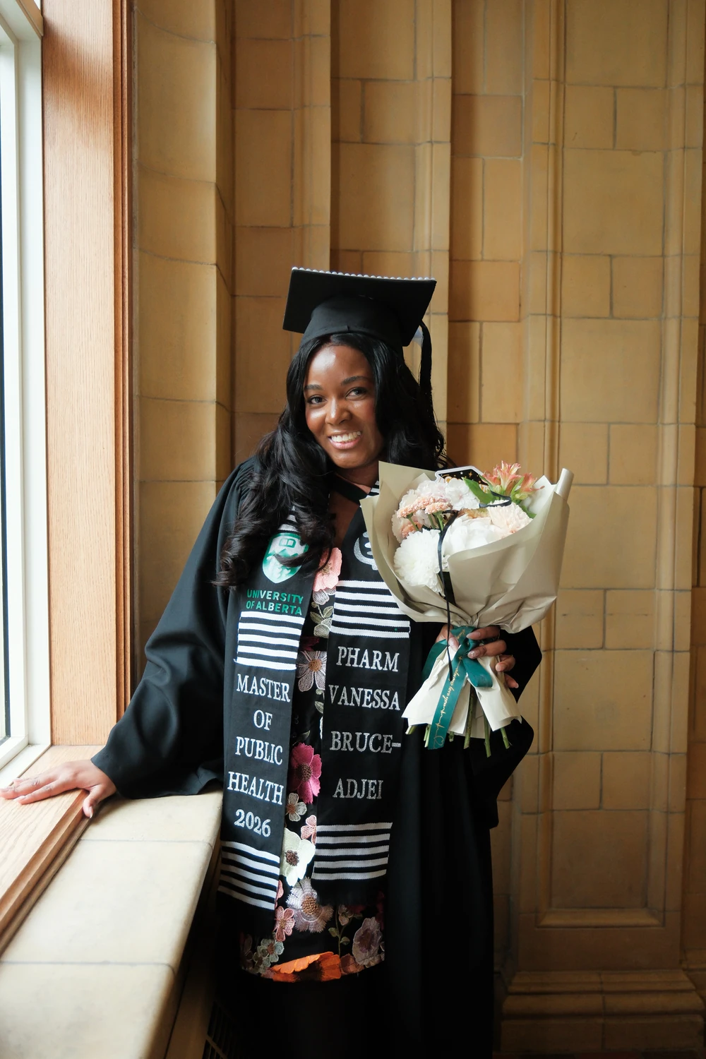 A smiling graduate holding a bouquet beside a tall window and warm stone wall.