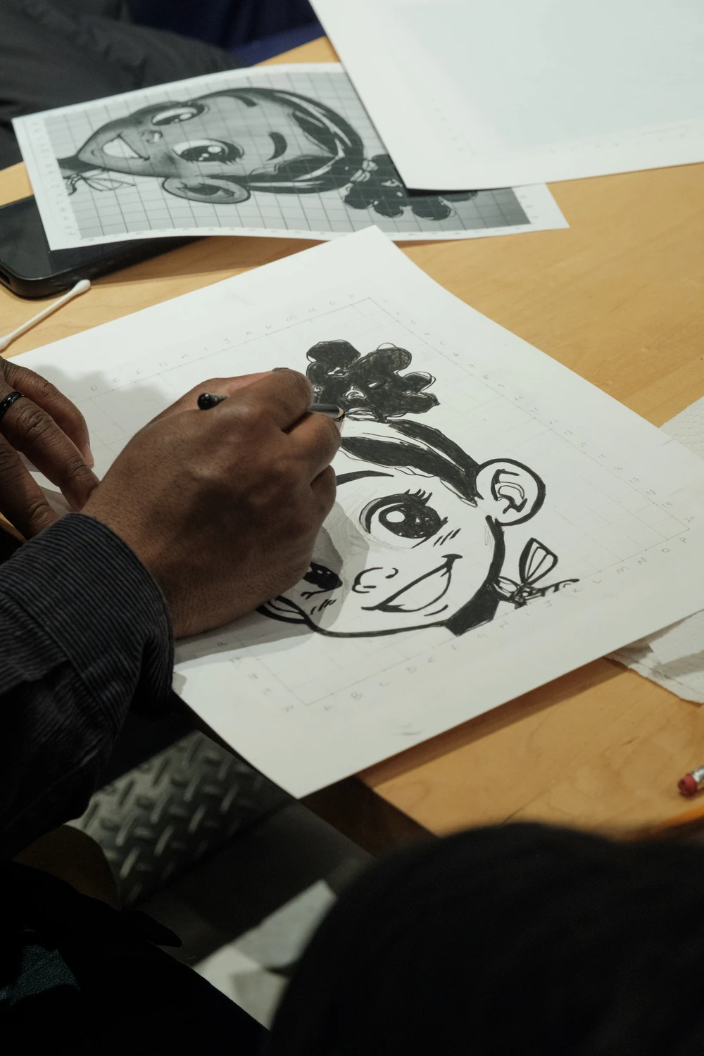 A hand drawing a character on paper beside a reference image at an Art with Ola workshop.