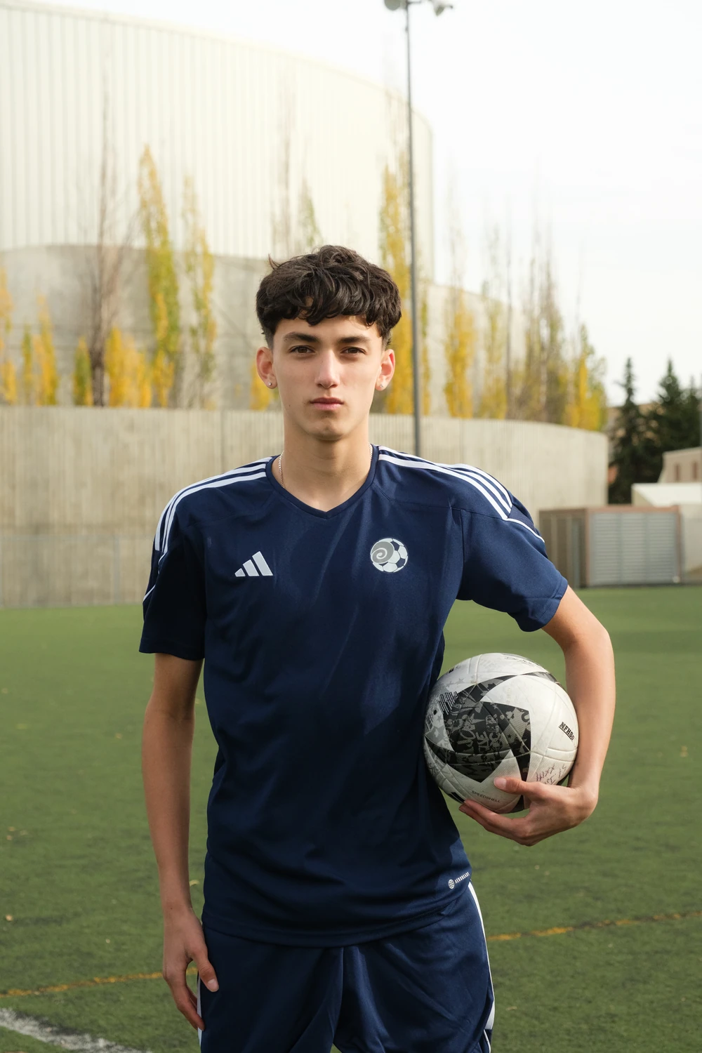 A football player standing on the pitch with a ball held beneath one arm.