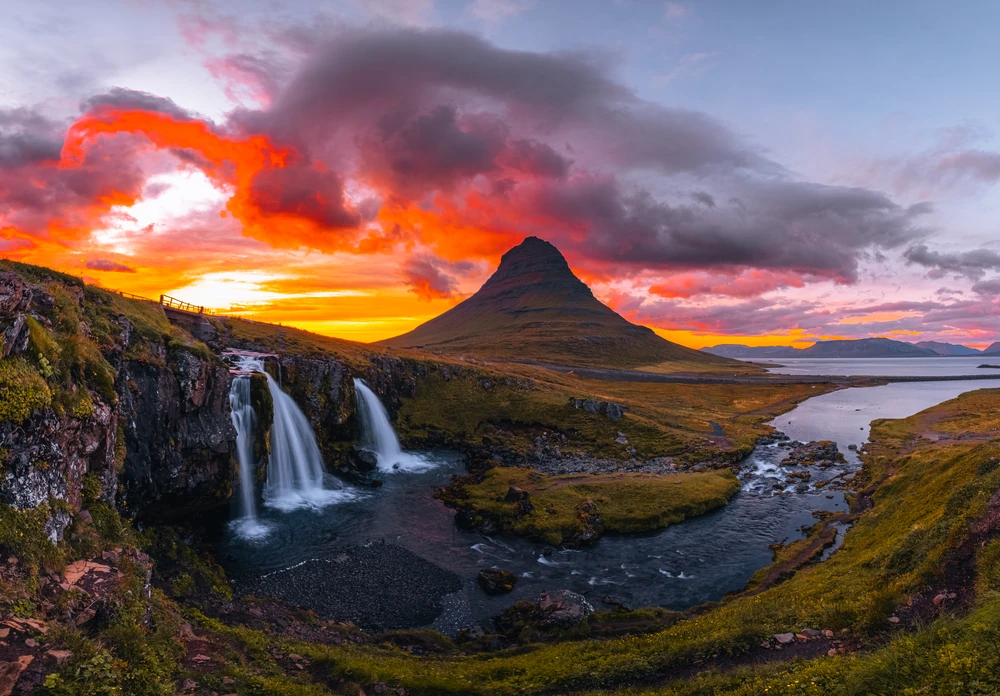 An Iceland panorama with waterfalls cascading into a winding river below a steep-sided mountain, under orange and pink sunset clouds.