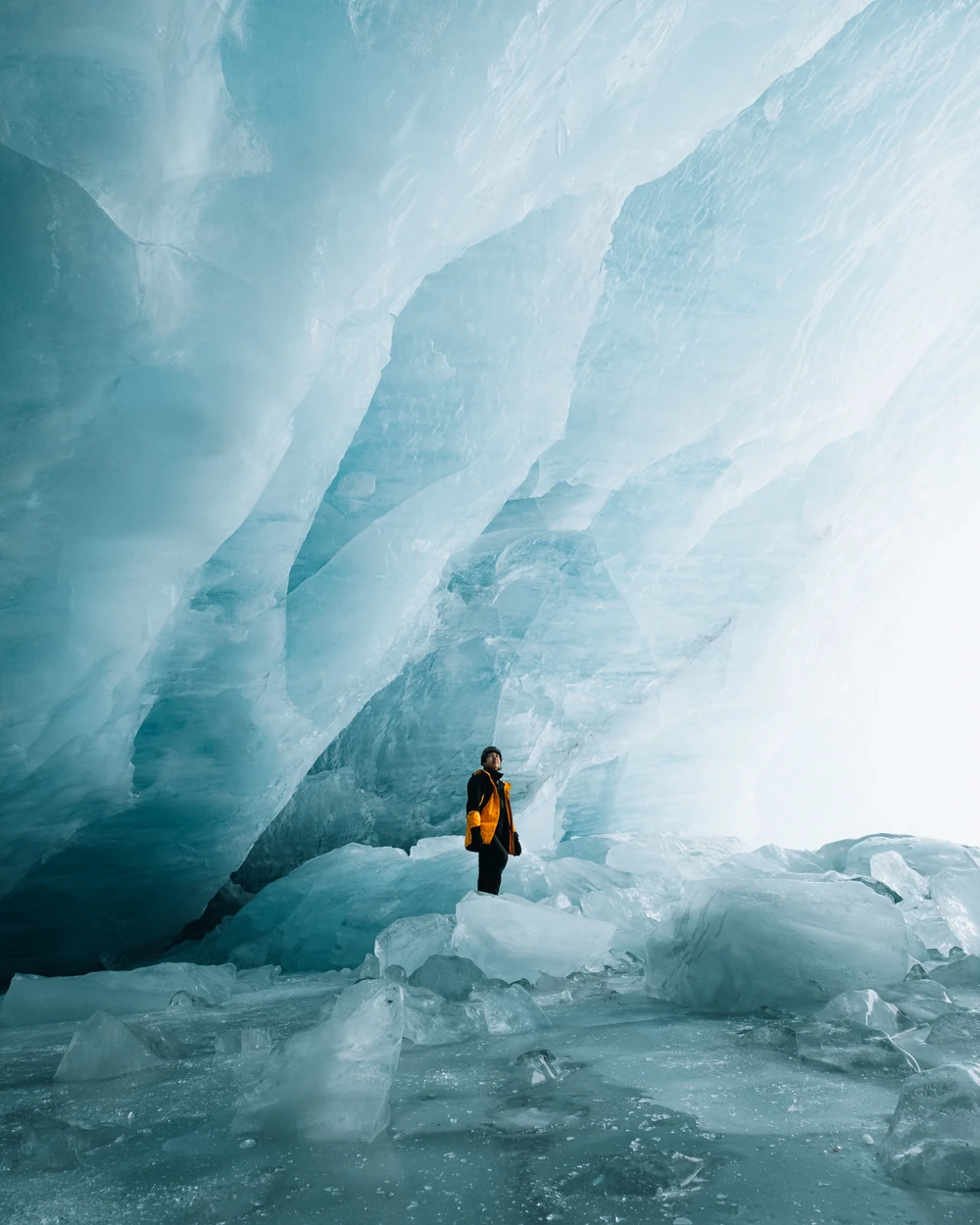 Aidan Lee in a yellow jacket standing beneath a towering blue glacier ice formation.