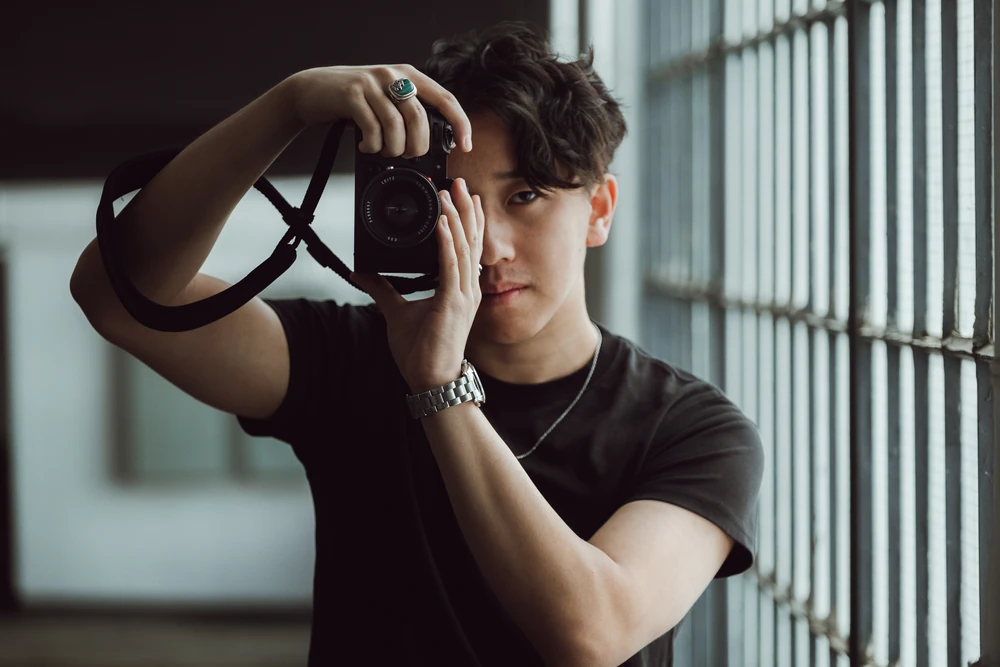 Aidan Lee holding a camera beside tall windows in a softly lit portrait.